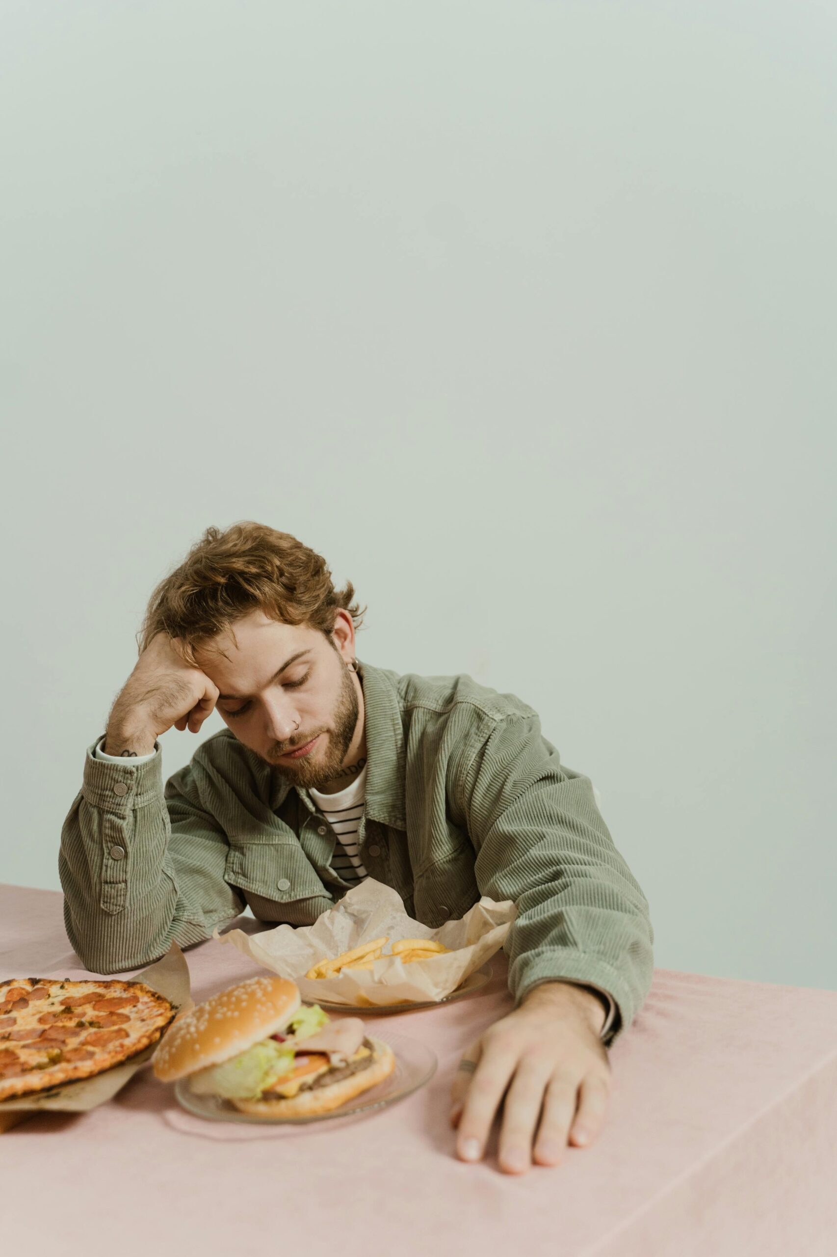 Young man appears tired with fast food burger and pizza on table in a relaxed setting.